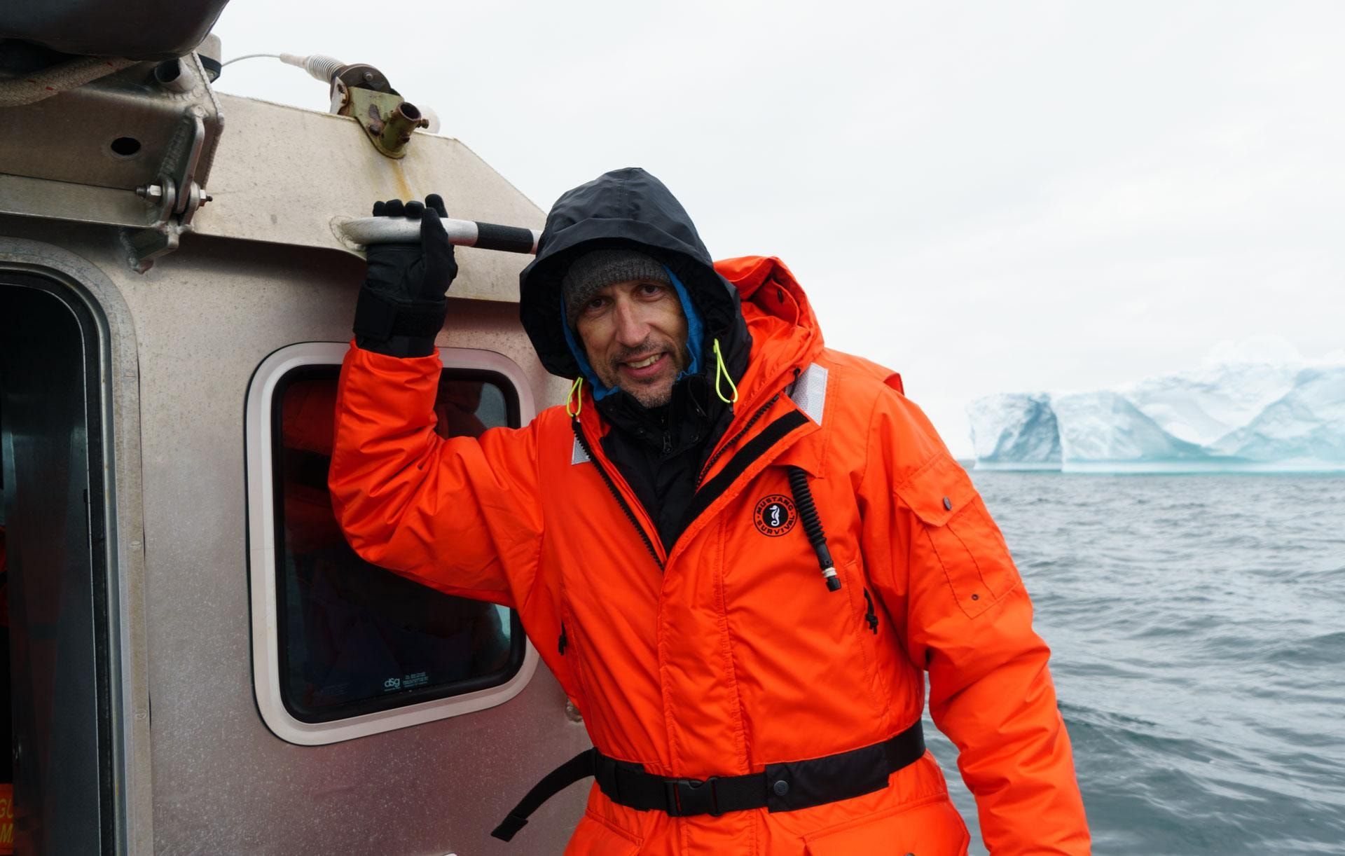 Person in orangefarbenem Überlebensanzug mit Kapuze hält sich an der Kabine eines Boots fest, im Hintergrund ein Eisberg auf dem Meer