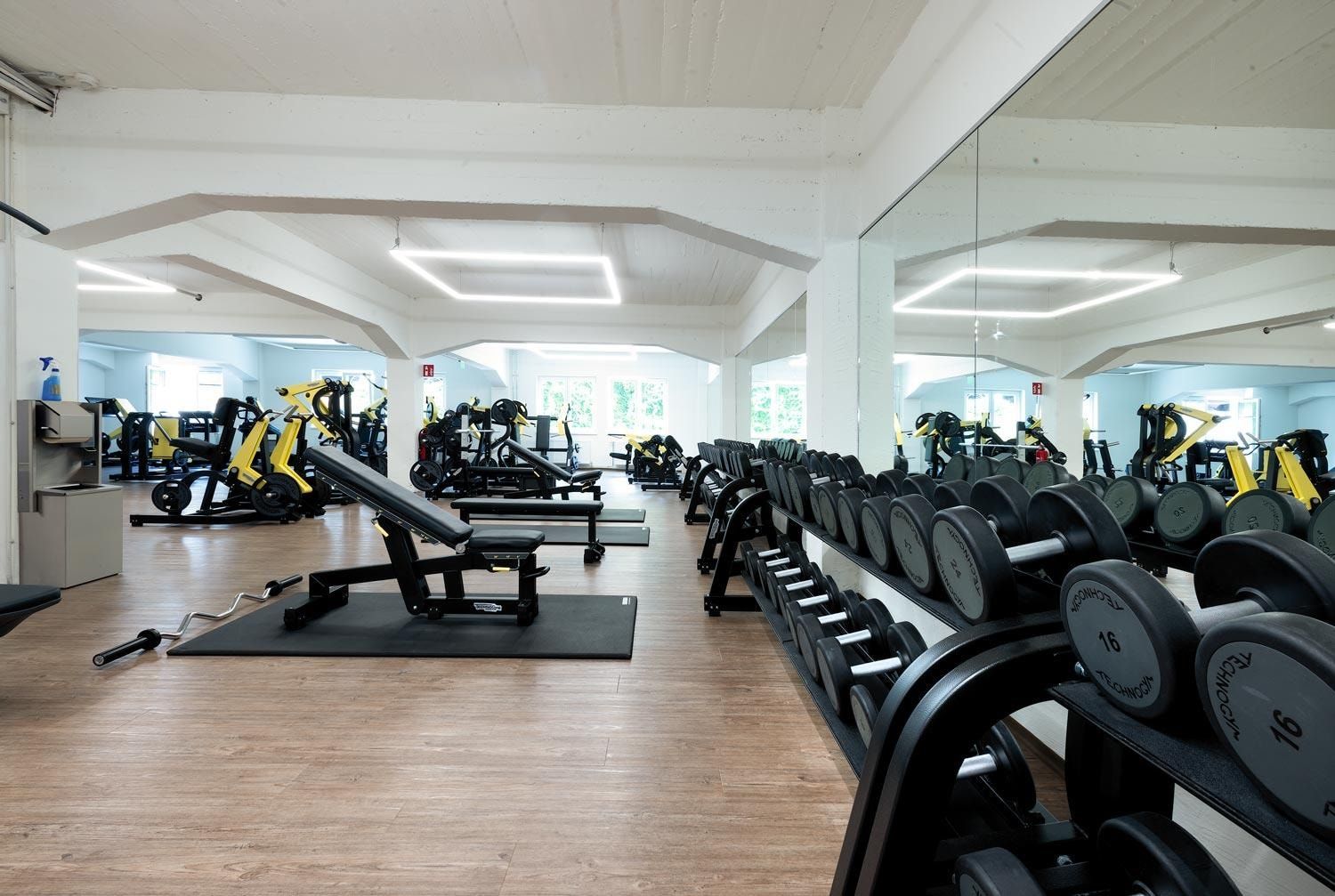Heller Fitnessraum mit Hantelständer, Kraftgeräten, Hantelbank und großer Spiegelwand