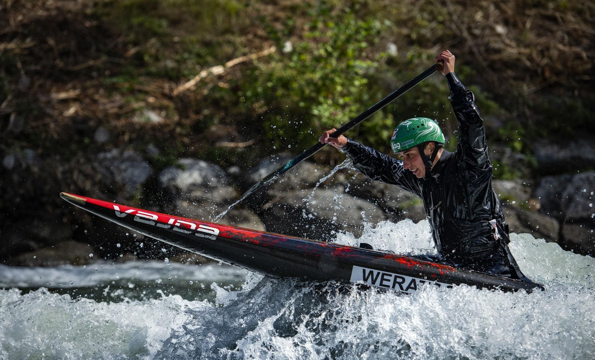 Person im Kanu mit grünem Helm im Wildwasser, das Paddel schräg nach oben gehalten, Gischt spritzt