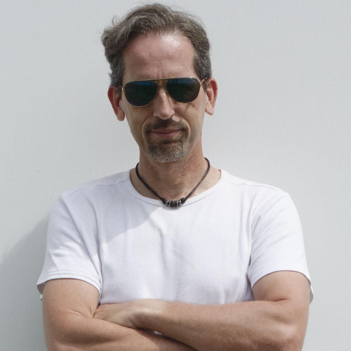 Portrait einer Person mit Sonnenbrille, Kinnbart und weißem T-Shirt, die Arme verschränkt, vor heller Wand