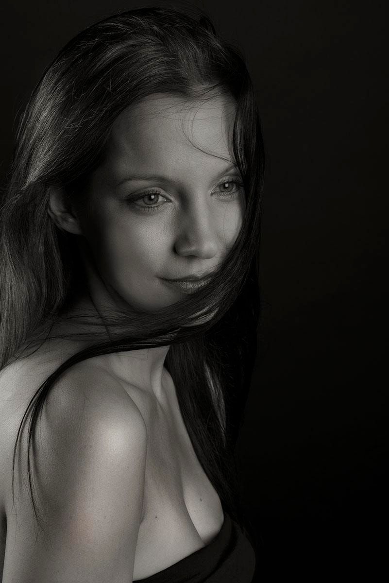 Schwarzweiß-Portrait mit langem dunklem Haar, das über Hals und Schulter fällt, gesenkter Blick zur Seite, dunkler Hintergrund