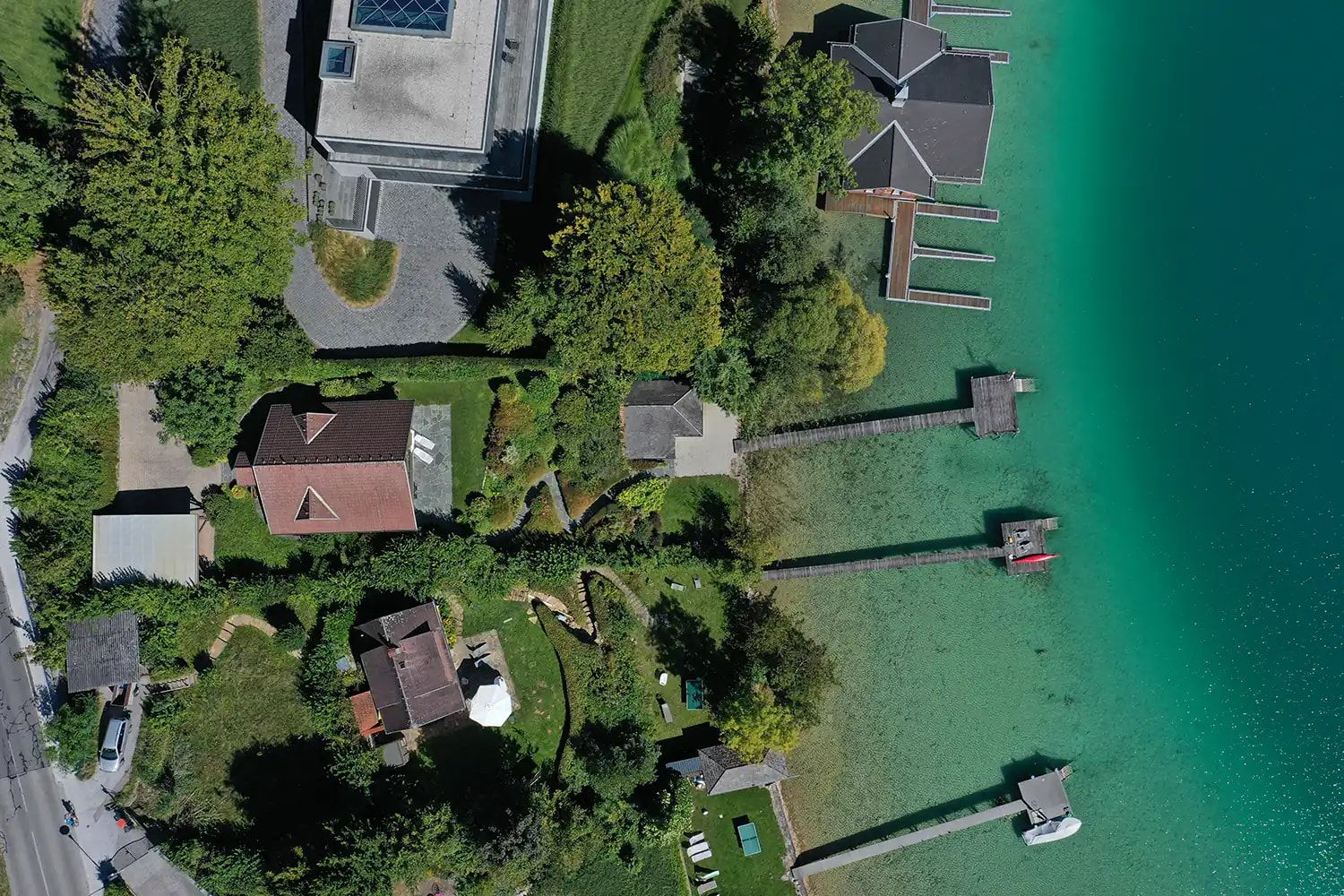 Senkrechter Blick aus der Luft auf ein Seeufer mit Häusern, Gärten, Holzstegen und türkisem Wasser