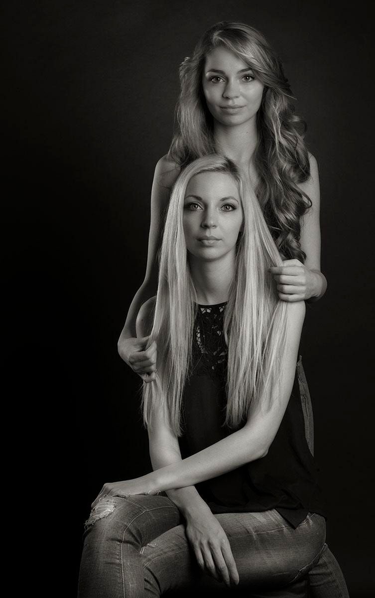 Schwarzweiß-Portrait zweier Personen mit langem Haar, eine sitzt, die andere steht dahinter und legt die Hände auf deren Schultern, dunkler Hintergrund